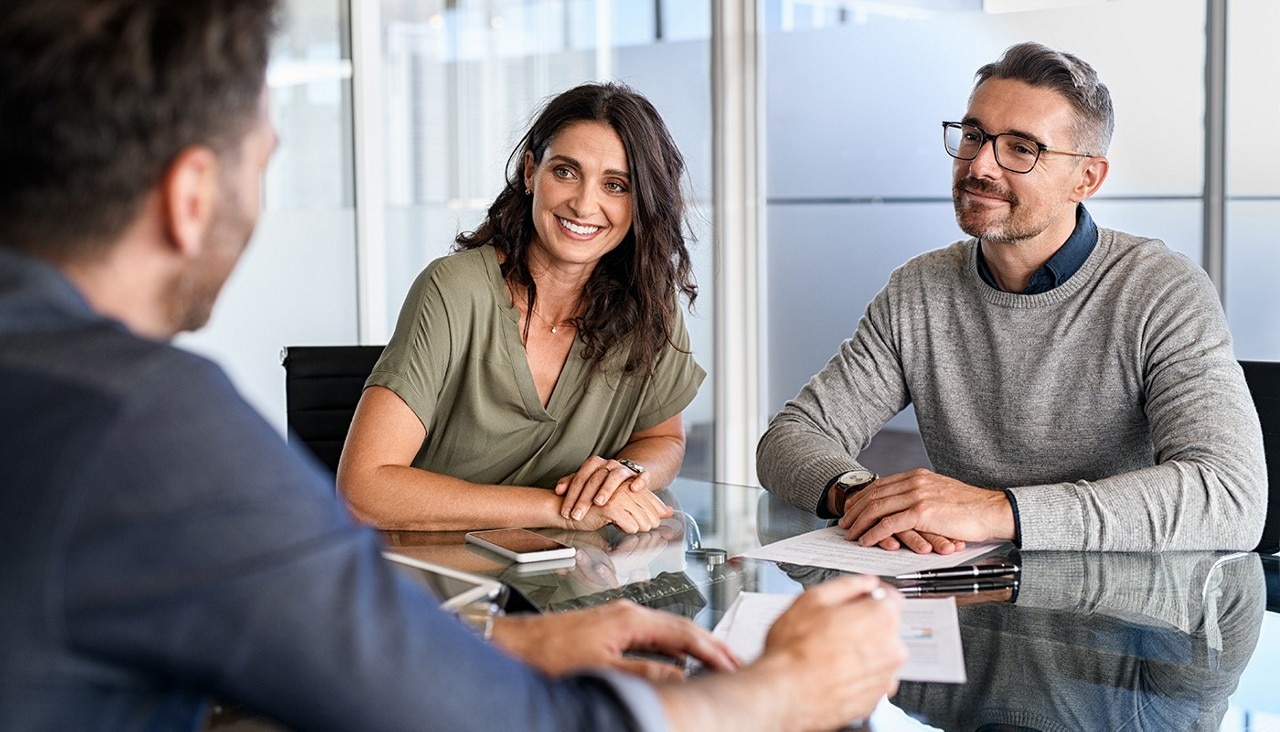 Smiling couple sitting at a table in an office as a life insurance agent speaks to them