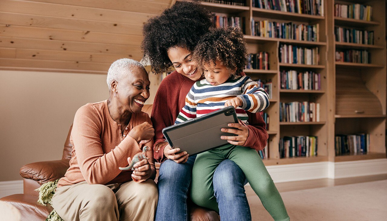 Grandmother, mother and child sit together smiling, looking at a tablet